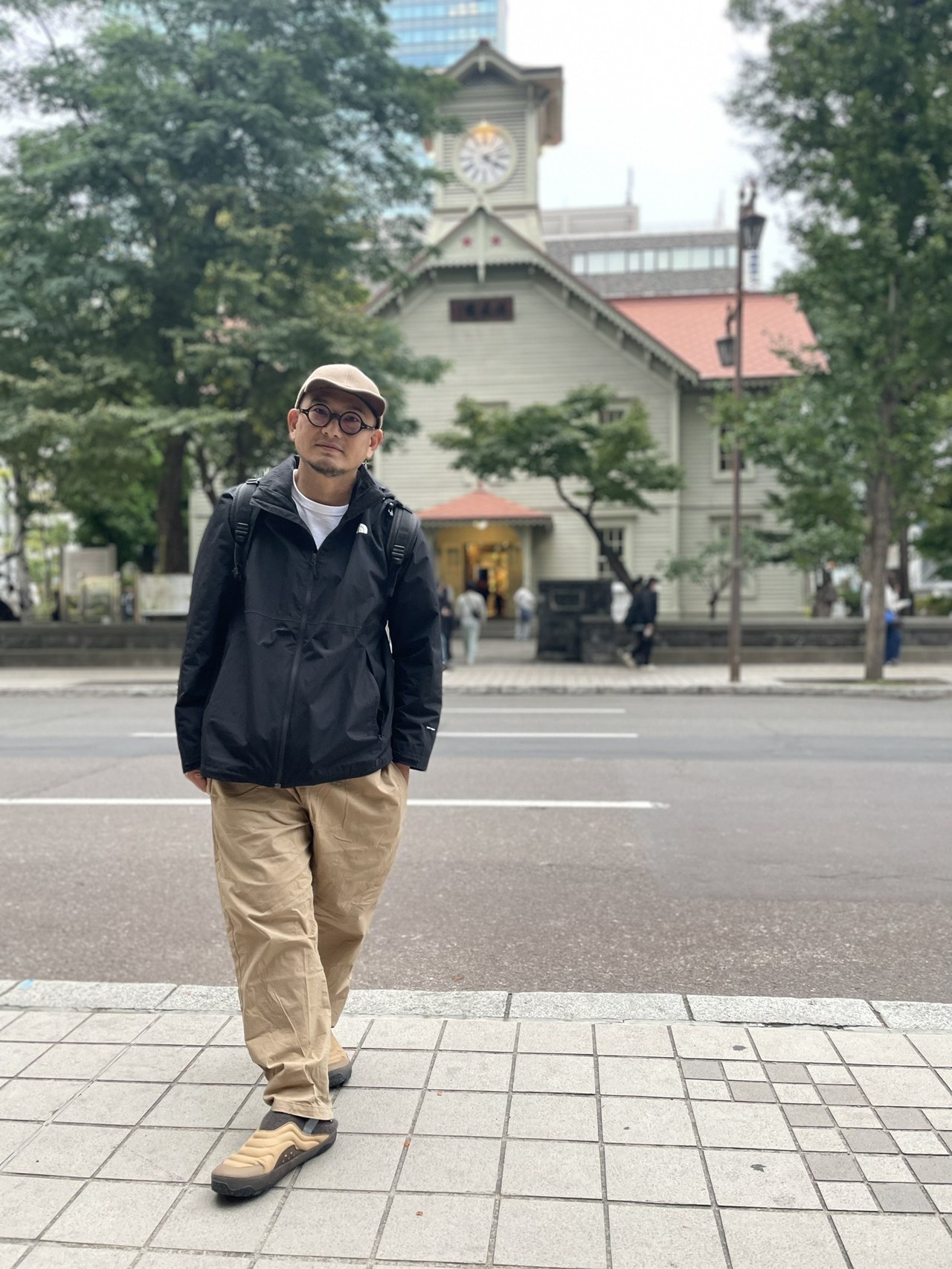 申廣堂主理人劉晉輔，於札幌時計台前的全身像：戴米色帽與圓框眼鏡、黑色外套與卡其長褲，背後為紅屋頂的木造鐘樓建築與行道樹