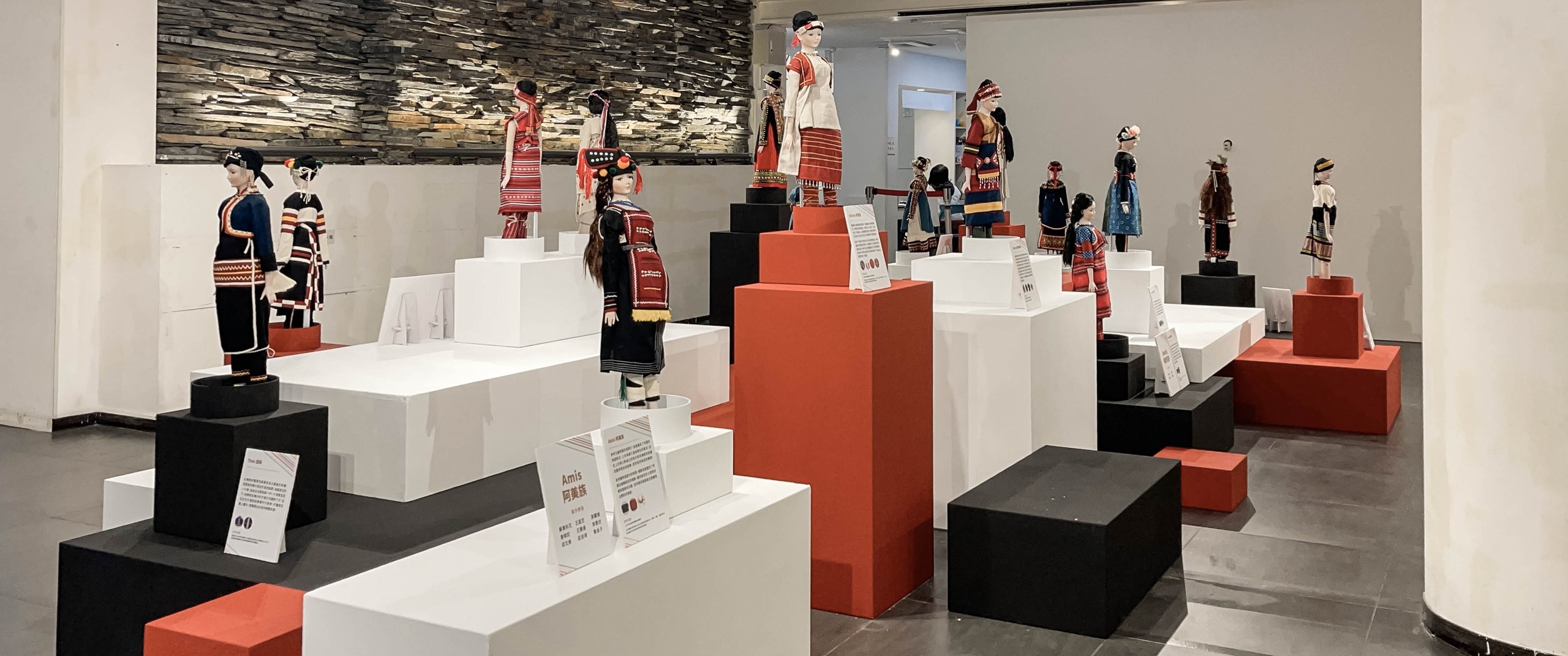 原住民族服飾展區：各族傳統服飾娃娃立於紅、白、黑三色高低錯落的塊體展台上，後方石牆與「阿美族 Amis」圖文說明卡，展台節奏明快而有秩序