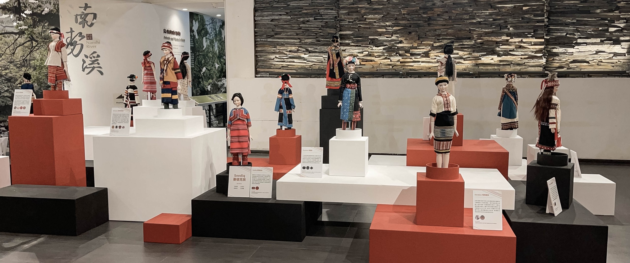 原住民族服飾展區全景：以「南勢溪 Nanshi River」為背景牆，賽德克、卑南、噶瑪蘭等各族服飾娃娃分立於紅白黑塊體展台，構成一條族群文化的動線