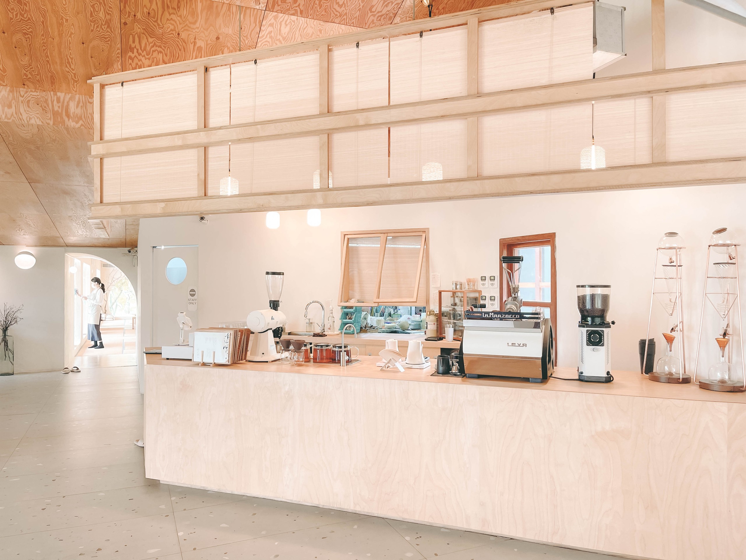 透明空氣吧台:淺色木夾板吧台與 La Marzocco 咖啡機、銅質手沖架,後方木格柵隔屏與圓窗,明亮自然光