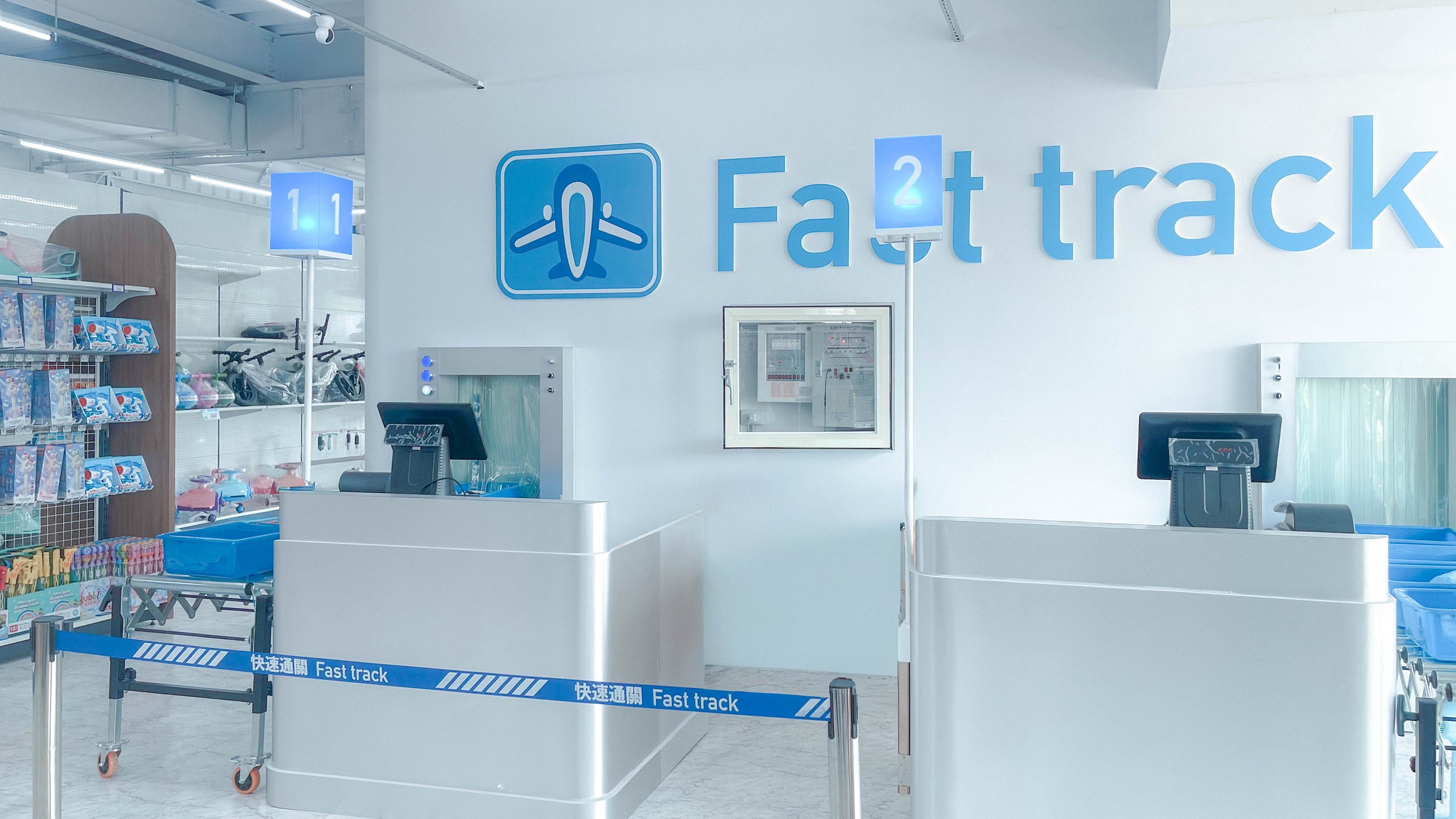 快速通關玩具店室內 Fast track 通關區:白牆上藍色飛機標誌與大字 Fast track,金屬結帳檯與藍色登機門號燈箱,像機場安檢通道