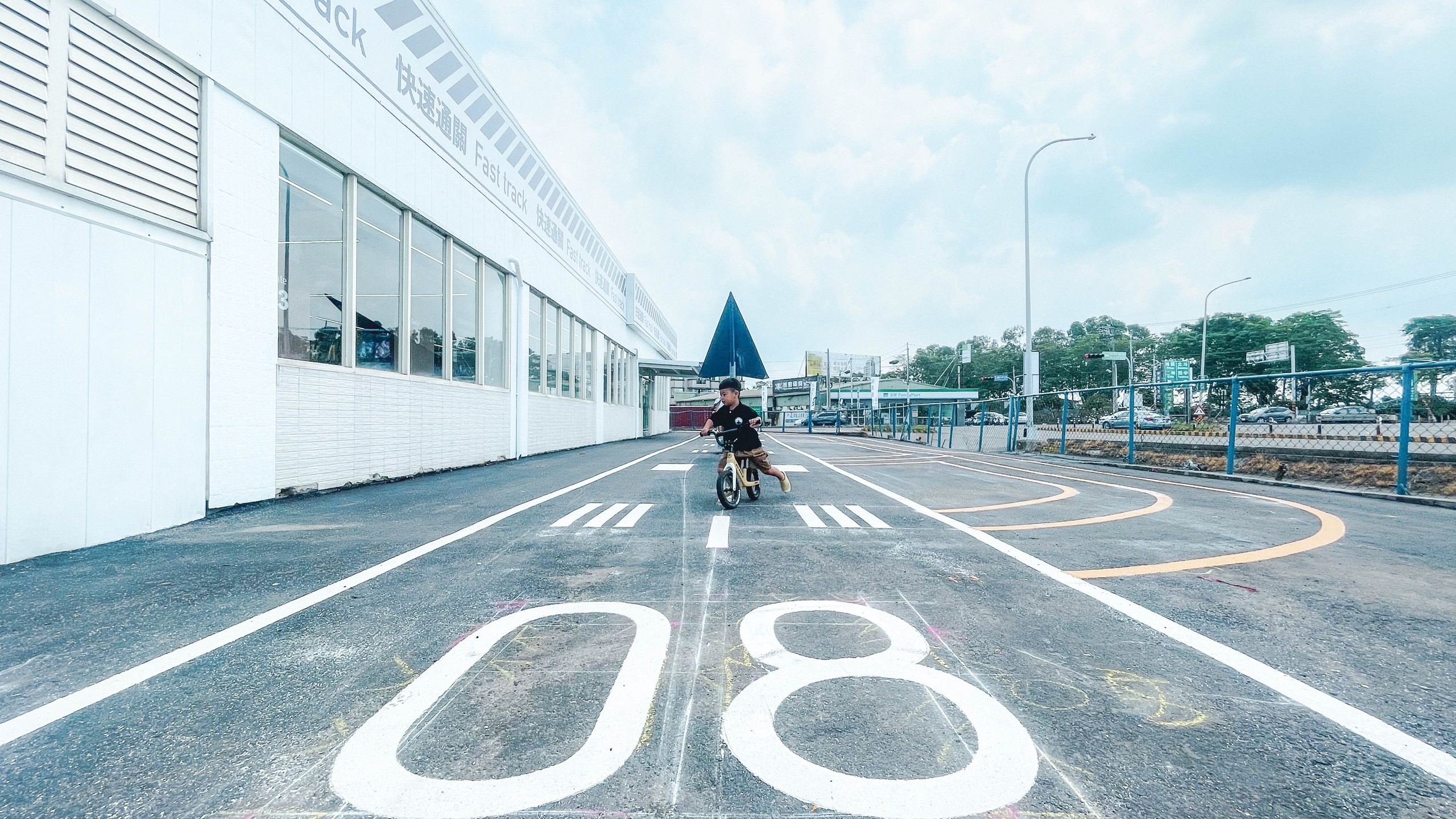 快速通關玩具店戶外跑道：柏油地面漆上白色跑道標線與大大的「08」編號，孩子騎著平衡車滑過斑馬線，建築側牆印著快速通關 Fast track